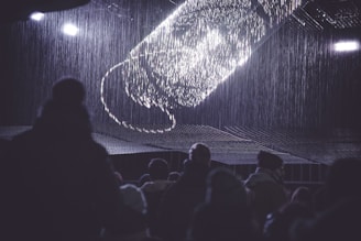 An audience is gathered in a dark environment, looking up at a digital or light installation in the shape of a large screw or twisting structure. The display emits bright, white light, creating a contrast against the surrounding darkness. Light seems to cascade down like rain, and silhouettes of people are visible in the foreground.