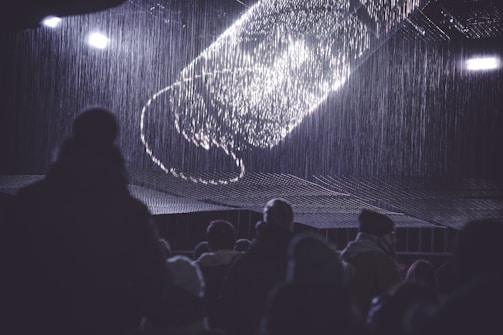 An audience is gathered in a dark environment, looking up at a digital or light installation in the shape of a large screw or twisting structure. The display emits bright, white light, creating a contrast against the surrounding darkness. Light seems to cascade down like rain, and silhouettes of people are visible in the foreground.