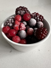 A white bowl filled with an assortment of frozen berries, including blackberries, raspberries, and cherries. A light layer of frost coats the berries, highlighting their texture and rich colors.