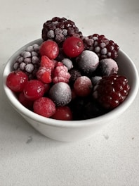 A white bowl filled with an assortment of frozen berries, including blackberries, raspberries, and cherries. A light layer of frost coats the berries, highlighting their texture and rich colors.