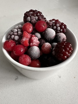 A white bowl filled with an assortment of frozen berries, including blackberries, raspberries, and cherries. A light layer of frost coats the berries, highlighting their texture and rich colors.