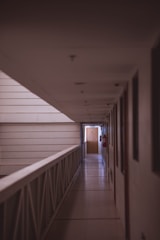 A dimly lit hallway with a tiled floor and a railing on the left side. Doors line the right side of the corridor, leading to rooms. A wooden door at the end is illuminated by soft natural light. Walls are neutral-colored, and there are a couple of red fire extinguishers mounted on the wall.