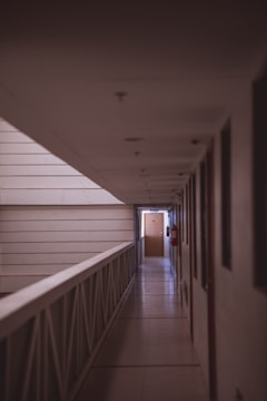 A dimly lit hallway with a tiled floor and a railing on the left side. Doors line the right side of the corridor, leading to rooms. A wooden door at the end is illuminated by soft natural light. Walls are neutral-colored, and there are a couple of red fire extinguishers mounted on the wall.