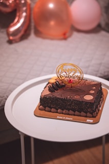 A square chocolate birthday cake with decorative toppings and a 'Happy Birthday' sign sits on a round white table. The cake is adorned with chocolate sprinkles and small mounds around the base. In the background, a quilted grey surface and several inflated balloons, including a number balloon, are visible.