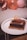 A square chocolate birthday cake with decorative toppings and a 'Happy Birthday' sign sits on a round white table. The cake is adorned with chocolate sprinkles and small mounds around the base. In the background, a quilted grey surface and several inflated balloons, including a number balloon, are visible.