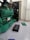 A large industrial generator with a green casing is situated indoors. Several black battery units connected by cables are placed on the floor beside the generator. The environment appears to be clean and organized, with a grey painted floor and white walls. Additional equipment is visible in the background, contributing to the machine's operation.