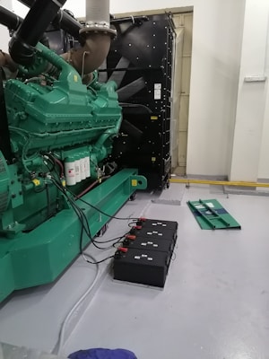 A large industrial generator with a green casing is situated indoors. Several black battery units connected by cables are placed on the floor beside the generator. The environment appears to be clean and organized, with a grey painted floor and white walls. Additional equipment is visible in the background, contributing to the machine's operation.