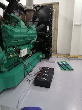 A large industrial generator with a green casing is situated indoors. Several black battery units connected by cables are placed on the floor beside the generator. The environment appears to be clean and organized, with a grey painted floor and white walls. Additional equipment is visible in the background, contributing to the machine's operation.