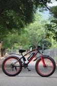 A black and red electric bicycle stands on a paved road. The bike features thick tires and a sturdy frame with branding that reads 'VOLTEBYK.' Surrounding the road are lush green trees, creating a peaceful and natural environment.
