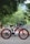 A black and red electric bicycle stands on a paved road. The bike features thick tires and a sturdy frame with branding that reads 'VOLTEBYK.' Surrounding the road are lush green trees, creating a peaceful and natural environment.