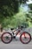 A black and red electric bicycle stands on a paved road. The bike features thick tires and a sturdy frame with branding that reads 'VOLTEBYK.' Surrounding the road are lush green trees, creating a peaceful and natural environment.