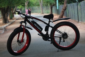 A mountain bike with thick tires is parked on a shaded pathway. The bike is predominantly black with red accents, including the rims and parts of the frame. There is greenery in the background, and two blurred figures are walking away on the path.