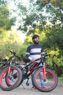 A person standing outdoors beside two electric bicycles with wide tires. The person wears a blue and gray striped sweater and jeans. The background features dense green foliage and a path.