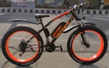 A black and orange electric bike with thick tires is positioned on a street, against the backdrop of an urban wall mural. The bike features a sturdy frame, wide orange-rimmed wheels, and fenders over both wheels.