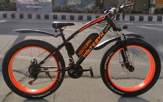 A black and orange electric bike with thick tires is positioned on a street, against the backdrop of an urban wall mural. The bike features a sturdy frame, wide orange-rimmed wheels, and fenders over both wheels.