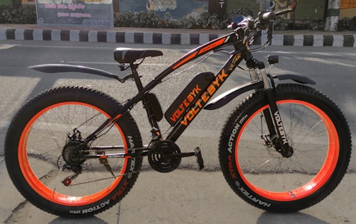 A black and orange electric bike with thick tires is positioned on a street, against the backdrop of an urban wall mural. The bike features a sturdy frame, wide orange-rimmed wheels, and fenders over both wheels.