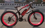 An electric bicycle with vibrant red rims and black frame sits on a road. The bike is well-equipped with fat tires, a cushioned seat, and front suspension. It features branding and model information on the frame. The background includes a painted mural and a road with traffic markings.