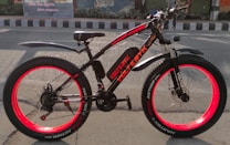 An electric bicycle with vibrant red rims and black frame sits on a road. The bike is well-equipped with fat tires, a cushioned seat, and front suspension. It features branding and model information on the frame. The background includes a painted mural and a road with traffic markings.