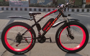 An electric bicycle with vibrant red rims and black frame sits on a road. The bike is well-equipped with fat tires, a cushioned seat, and front suspension. It features branding and model information on the frame. The background includes a painted mural and a road with traffic markings.