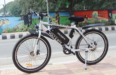 A white electric bicycle is parked on a tiled sidewalk beside a road. The bicycle is branded with 'VOLT E BYK' and features a black saddle and tires. In the background, a colorful mural painted on a wall depicts various artistic designs and scenes, with trees and greenery nearby.
