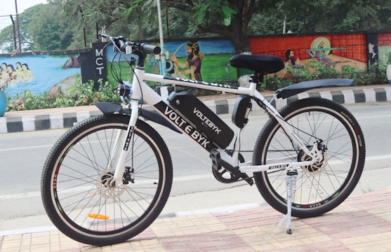 A white electric bicycle is parked on a tiled sidewalk beside a road. The bicycle is branded with 'VOLT E BYK' and features a black saddle and tires. In the background, a colorful mural painted on a wall depicts various artistic designs and scenes, with trees and greenery nearby.