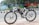 A white electric bicycle is parked on a tiled sidewalk beside a road. The bicycle is branded with 'VOLT E BYK' and features a black saddle and tires. In the background, a colorful mural painted on a wall depicts various artistic designs and scenes, with trees and greenery nearby.
