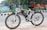 A white electric bicycle is parked on a tiled sidewalk beside a road. The bicycle is branded with 'VOLT E BYK' and features a black saddle and tires. In the background, a colorful mural painted on a wall depicts various artistic designs and scenes, with trees and greenery nearby.