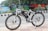 A white electric bicycle is parked on a tiled sidewalk beside a road. The bicycle is branded with 'VOLT E BYK' and features a black saddle and tires. In the background, a colorful mural painted on a wall depicts various artistic designs and scenes, with trees and greenery nearby.