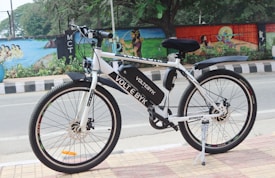 A white electric bicycle is parked on a tiled sidewalk beside a road. The bicycle is branded with 'VOLT E BYK' and features a black saddle and tires. In the background, a colorful mural painted on a wall depicts various artistic designs and scenes, with trees and greenery nearby.
