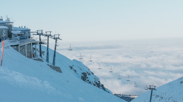 A snowy mountain landscape featuring a ski lift and a station at the edge of a slope. The sky is clear with a layer of clouds visible below, giving a sense of height and vastness.