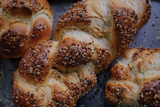 Freshly baked braided loaves of bread topped with a generous sprinkling of sesame and poppy seeds, resting on a dark baking tray.