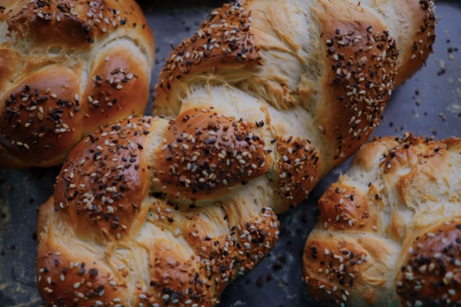 Freshly baked braided loaves of bread topped with a generous sprinkling of sesame and poppy seeds, resting on a dark baking tray.