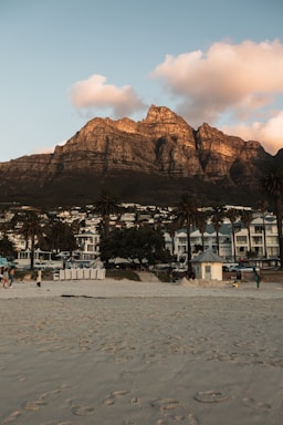 A serene beachfront scene is depicted, showing sandy beach in the foreground with a few people walking. Behind the beach, there are tall palm trees, residential buildings, and houses with a backdrop of rugged, rocky mountains. The sunlight casts a warm glow on the mountains and clouds hang in the sky.