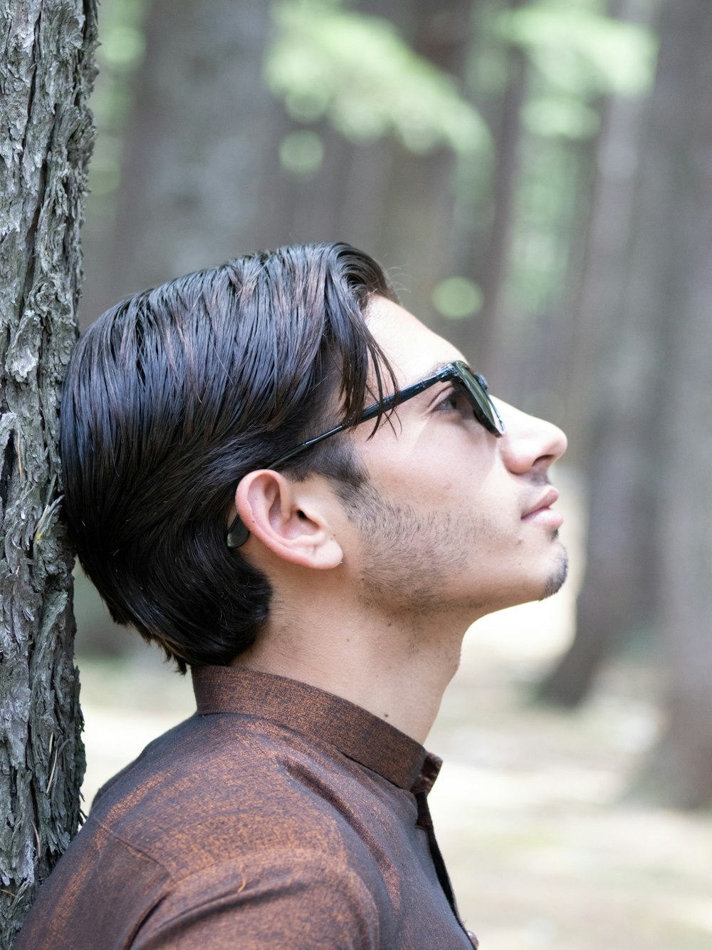 a man with black hair and glasses leaning against a tree