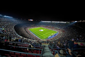 A large, illuminated football stadium filled with spectators. The green soccer field is centrally located, with players visible near the center circle. The stands are packed with people, creating a vibrant and dynamic atmosphere. Stadium lights shine down onto the field, contrasting with the dark night sky.
