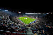 A large, illuminated football stadium filled with spectators. The green soccer field is centrally located, with players visible near the center circle. The stands are packed with people, creating a vibrant and dynamic atmosphere. Stadium lights shine down onto the field, contrasting with the dark night sky.