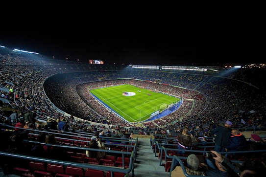 A large, illuminated football stadium filled with spectators. The green soccer field is centrally located, with players visible near the center circle. The stands are packed with people, creating a vibrant and dynamic atmosphere. Stadium lights shine down onto the field, contrasting with the dark night sky.