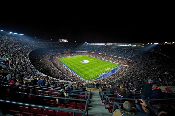 A large, illuminated football stadium filled with spectators. The green soccer field is centrally located, with players visible near the center circle. The stands are packed with people, creating a vibrant and dynamic atmosphere. Stadium lights shine down onto the field, contrasting with the dark night sky.