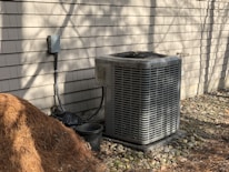 An outdoor air conditioning unit is positioned against a beige shingled wall. There is a pile of pine straw mulch on the ground nearby, along with a small black plastic bucket. The area is surrounded by small stones, and the scene is bathed in natural sunlight, casting shadows against the wall.