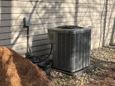 An outdoor air conditioning unit is positioned against a beige shingled wall. There is a pile of pine straw mulch on the ground nearby, along with a small black plastic bucket. The area is surrounded by small stones, and the scene is bathed in natural sunlight, casting shadows against the wall.