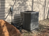 An outdoor air conditioning unit is positioned against a beige shingled wall. There is a pile of pine straw mulch on the ground nearby, along with a small black plastic bucket. The area is surrounded by small stones, and the scene is bathed in natural sunlight, casting shadows against the wall.