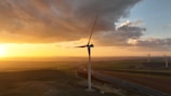 A large wind turbine stands prominently against a backdrop of a vivid sunset, with the sky displaying a blend of orange and yellow hues. The surrounding landscape consists of open fields and several other wind turbines in the distance, with a highway running through the scene.