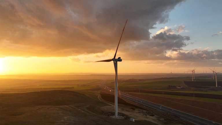 A large wind turbine stands prominently against a backdrop of a vivid sunset, with the sky displaying a blend of orange and yellow hues. The surrounding landscape consists of open fields and several other wind turbines in the distance, with a highway running through the scene.