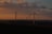 Wind turbines are scattered across a landscape under a dramatic sky with rich clouds and warm sunset lighting. The horizon features rolling hills, and the turbines are positioned both in silhouette and with sunlight catching parts of the blades.