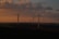 Wind turbines are scattered across a landscape under a dramatic sky with rich clouds and warm sunset lighting. The horizon features rolling hills, and the turbines are positioned both in silhouette and with sunlight catching parts of the blades.