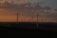 Wind turbines are scattered across a landscape under a dramatic sky with rich clouds and warm sunset lighting. The horizon features rolling hills, and the turbines are positioned both in silhouette and with sunlight catching parts of the blades.