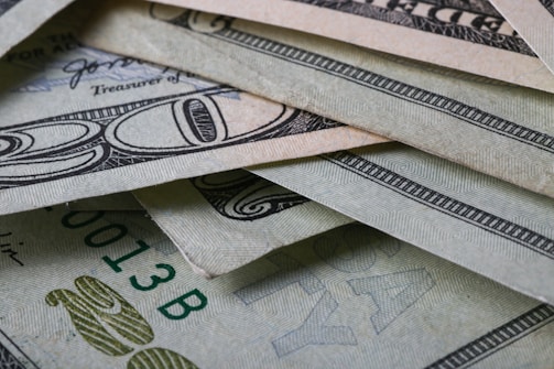 A close-up view of multiple U.S. dollar bills arranged in an overlapping manner. The intricate details of the currency's security features and patterns are visible, showing shades of green and grey.