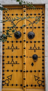 A large, ornately decorated wooden door painted in a vibrant yellow, featuring intricate black patterns and studded designs. The door has two large circular metal knockers and is framed by a light stone structure. Overhanging green leaves add a natural touch to the scene.
