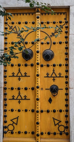 A large, ornately decorated wooden door painted in a vibrant yellow, featuring intricate black patterns and studded designs. The door has two large circular metal knockers and is framed by a light stone structure. Overhanging green leaves add a natural touch to the scene.