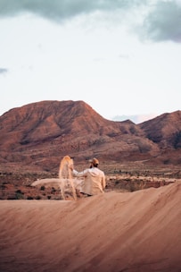 A person in casual clothing and a cap appears to be seated on a sand dune, sifting sand through their hand. The background features rugged, rocky mountains with a largely clear sky.